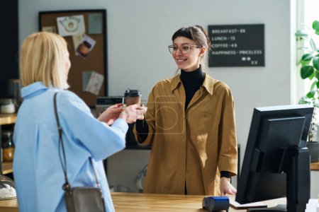 Téléchargez les photos : Jeune serveuse en lunettes regardant blonde mature cliente avec le sourire tout en passant sa tasse de café sur le comptoir dans le café - en image libre de droit