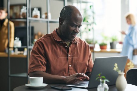 Téléchargez les photos : Homme d'affaires afro-américain mature tapant sur le clavier de l'ordinateur portable tout en étant assis à table dans un café, ayant une tasse de café et de réseautage - en image libre de droit