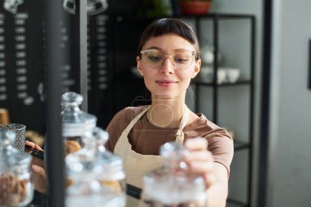 Téléchargez les photos : Jeune employée souriante vérifiant les restes de tisane sèche dans de petits pots debout sur une étagère tout en faisant la révision des produits dans le café - en image libre de droit