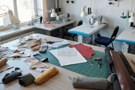 Workplace of tanner with sketches on papers, rolled leather textile, handtools and pieces of fabric against desks with sewing equipment