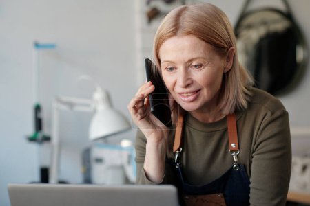 Téléchargez les photos : Femme mûre blonde souriante avec smartphone par oreille regardant l'écran de l'ordinateur portable et parlant à l'un des clients tout en spécifiant les détails de la commande - en image libre de droit