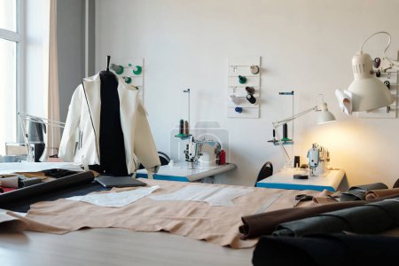 Parte de un amplio taller de cuero con equipo de costura, maniquí con chaqueta de cuero blanco y mesa con textil