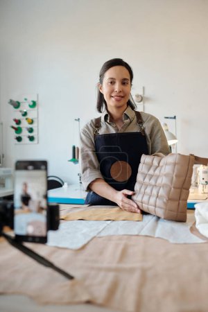 Téléchargez les photos : Jeune femme créative souriante dans un tablier avec un nouveau sac en cuir fait main regardant la caméra du smartphone tout en donnant masterclass en studio - en image libre de droit