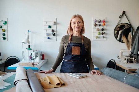 Smiling mature craftswoman in apron looking at camera in workshop or studio while standing by table with various leather and suede textile
