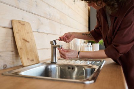 Téléchargez les photos : Mains de jeune femme au foyer en chemise marron essayant d'ouvrir le robinet métallique sur l'évier tout en restant debout près du comptoir en bois dans la cuisine à la maison - en image libre de droit