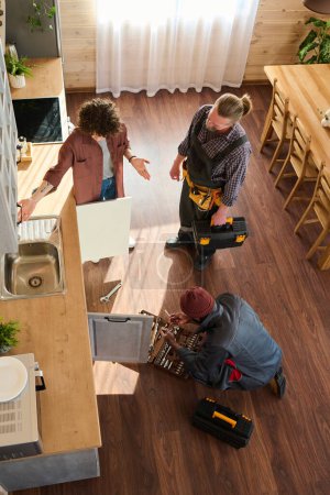 Téléchargez les photos : Vue ci-dessus de deux réparateurs en vêtements de travail vérifiant les tuyaux dans la cuisine tandis que l'un d'eux communiquant avec une femme au foyer troublée - en image libre de droit