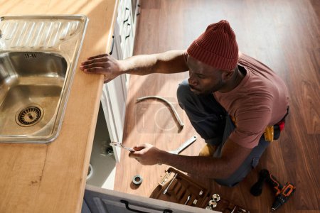 Téléchargez les photos : Angle élevé de jeune plombier afro-américain avec outil à main vérifiant les tuyaux à l'intérieur de l'armoire du comptoir de cuisine tout en se tenant sur les squats - en image libre de droit