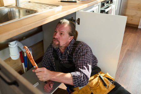 Téléchargez les photos : Réparateur barbu mature avec outil à main essayant de détecter le problème technique avec tuyau sous l'évier tout en travaillant dans l'armoire de cuisine - en image libre de droit
