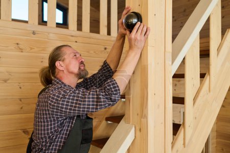 Photo for Bearded mature professional adjuster in workwear fixing alarm bell on wooden pile while standing on staircase in living room of apartment - Royalty Free Image