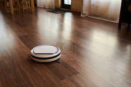 Téléchargez les photos : Partie de la cuisine spacieuse ou salon avec robot aspirateur se déplaçant sur le sol avec linoléum brun imitant stratifié en bois - en image libre de droit