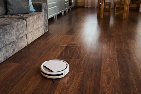 Téléchargez les photos : Partie de la cuisine spacieuse ou salon avec aspirateur robot se déplaçant le long du sol et le nettoyage linoléum imitant stratifié - en image libre de droit