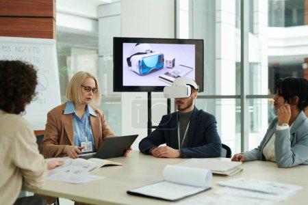 Photo for Young man with vr headset sitting by table among colleagues during presentation at meeting while mature woman using laptop - Royalty Free Image