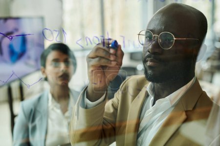 Photo for Focus on young serious African American businessman or coach with blue highlighter writing on transparent board during presentation - Royalty Free Image