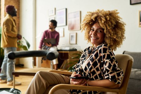 Téléchargez les photos : Jeune dirigeante afro-américaine réussie en tenue décontractée intelligente assise dans un fauteuil et regardant la caméra avec le sourire - en image libre de droit