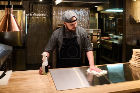 Téléchargez les photos : Jeune chef en uniforme debout près d'une grande table en bois et surface de nettoyage du panneau de poêle électrique avant de cuisiner une pizza pour les clients - en image libre de droit
