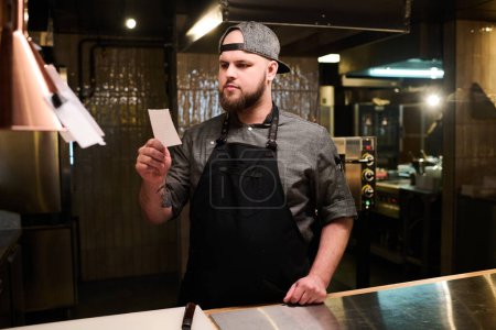 Téléchargez les photos : Jeune chef sérieux homme lisant des notes de recette sur petit papier tout en restant au travail dans la cuisine du restaurant ou de la pizzeria - en image libre de droit