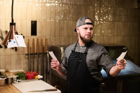 Téléchargez les photos : Jeune chef confiant du restaurant tenant de petits papiers avec des recettes tout en restant près du lieu de travail dans la cuisine et en choisissant l'un d'eux - en image libre de droit