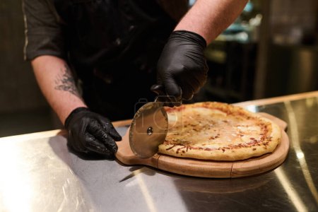 Photo for Hands in black rubber gloves of young chef cutting appetizing baked pizza with sharp knife on wooden board while standing by table in the kitchen - Royalty Free Image