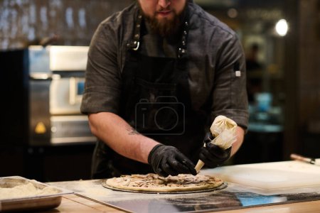 Téléchargez les photos : Mains dans les gants noirs du jeune chef pressant mélange écrasé d'ingrédients du sac à pâtisserie sur pain plat pendant la cuisson de la pizza - en image libre de droit