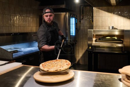 Foto de Joven chef poniendo pizza italiana horneada caliente sobre una mesa redonda de madera sobre una mesa grande mientras está de pie contra el horno eléctrico en la cocina - Imagen libre de derechos