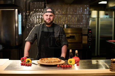 Téléchargez les photos : Joyeux jeune chef prospère et propriétaire d'un restaurant ou d'une pizzeria regardant la caméra tout en restant à table avec une pizza appétissante dans la cuisine - en image libre de droit