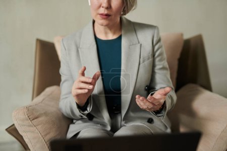 Foto de Primer plano del psicólogo confiado en traje gris sentado en sillón delante de la computadora portátil durante la sesión en línea y dando consejos al paciente - Imagen libre de derechos