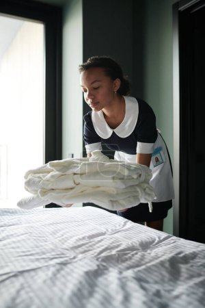 Foto de Camarera joven en uniforme que pone una pila de sábanas, toallas o albornoces doblados limpios en la cama mientras prepara la habitación de hotel para los huéspedes - Imagen libre de derechos