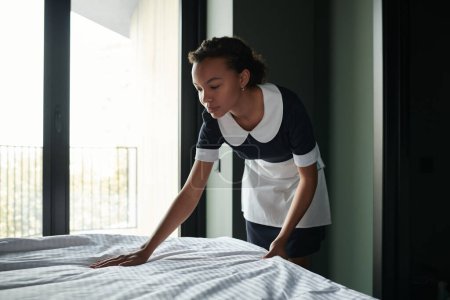 Photo for Side view of young housemaid putting clean bedclothes on bed while preparing room of luxurious five star hotel for new guests - Royalty Free Image