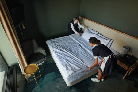 Photo for Above angle of young chambermaid and manager preparing double bed for guests while changing bedclothes before arrival of travelers - Royalty Free Image