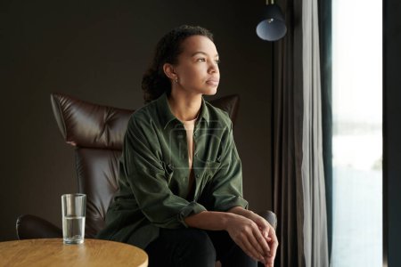Téléchargez les photos : Jeune femme afro-américaine malheureuse assise devant une grande fenêtre et regardant à travers elle tout en visitant le bureau de psychothérapeute - en image libre de droit