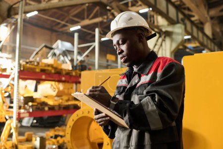 Photo for Serious young black man in workwear making technical notes in document while standing in front of camera against huge industrial machine - Royalty Free Image