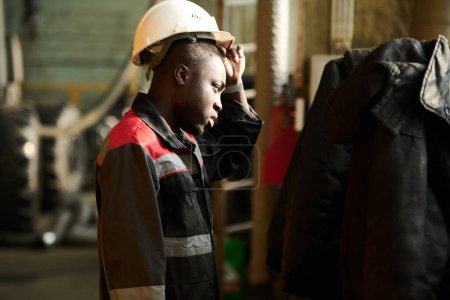 Téléchargez les photos : Jeune ingénieur afro-américain fatigué de l'usine industrielle enlevant le casque de protection après une dure journée de travail ou pendant une pause - en image libre de droit