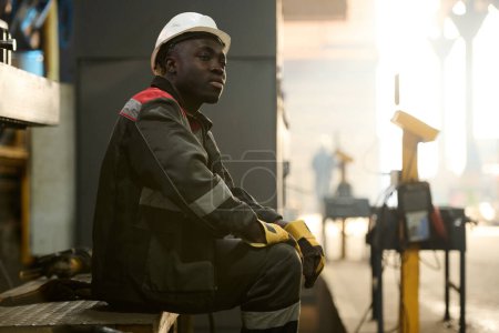Photo for Young tired African American workman sitting on metallic bench while resting at break in the middle of working day in factory workshop - Royalty Free Image
