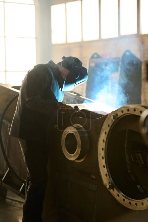 Foto de Vista lateral del ingeniero en máscara protectora y ropa de trabajo doblando sobre una enorme máquina industrial en el taller durante el proceso de soldadura por arco - Imagen libre de derechos
