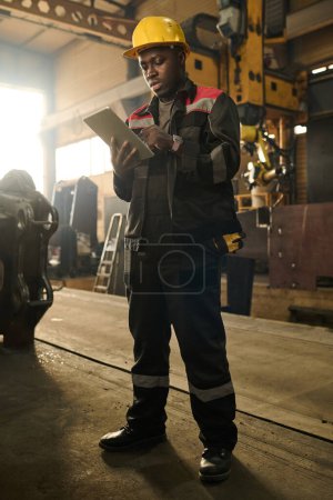 Foto de Joven afroamericano técnico masculino mirando a través de manual en línea mientras está de pie delante de la cámara y preparándose para la reparación de la máquina - Imagen libre de derechos