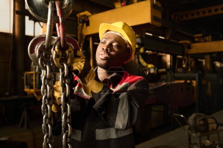 Photo for Young African American technician holding thick metallic chains and looking upwards while preparing for exploitation of machine - Royalty Free Image