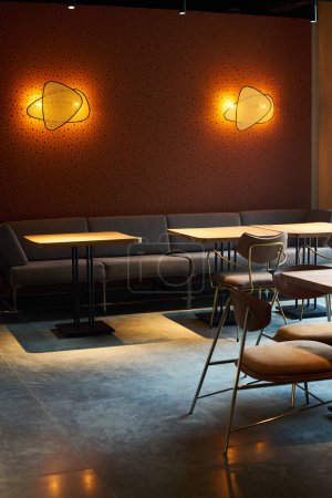 Foto de Parte de acogedora cafetería o salón en la planta baja del moderno hotel o centro de negocios con sofá a lo largo de la pared con dos lámparas, mesas y sillas - Imagen libre de derechos