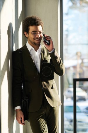 Photo for Young smiling multi-ethnic white collar worker with handbag on shoulders talking on smartphone while standing by wall in sunlit lounge - Royalty Free Image
