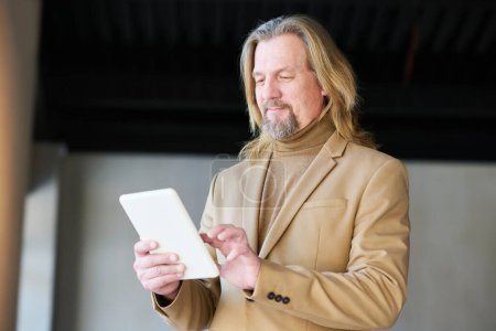 Téléchargez les photos : Homme d'affaires mature élégant en coffrage beige en utilisant une tablette tout en analysant les données graphiques en ligne ou en entrant des points de nouveau projet - en image libre de droit