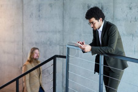 Foto de Joven hombre de negocios serio mirando a través de los datos en línea en la tableta, mientras que de pie contra barandilla en la escalera en el pasillo del centro de oficina - Imagen libre de derechos