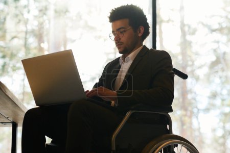 Foto de Joven hombre de negocios multiétnico con discapacidad utilizando el ordenador portátil mientras está sentado en silla de ruedas delante de la cámara y preparando un nuevo proyecto - Imagen libre de derechos