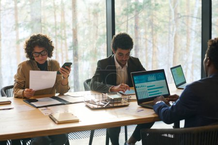 Junge interkulturelle Mitarbeiter sitzen im Sitzungssaal am Tisch, vernetzen sich, blättern in Papierdokumenten, nutzen Tablets und analysieren Daten
