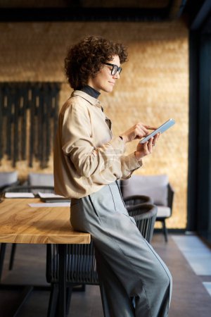 Photo for Side view of young confident female analyst in quiet luxury attire pointing at tablet screen while standing against wooden table in boardroom - Royalty Free Image