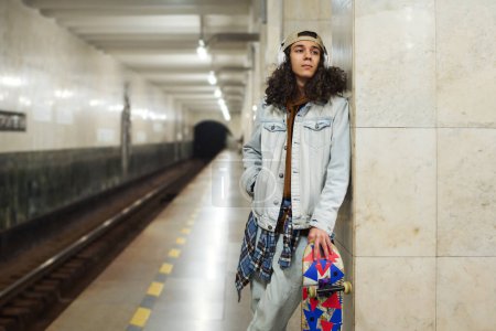 Foto de Elegante adolescente en ropa de casualwear y auriculares de pie junto a la columna de mármol en la estación de metro mientras espera el tren subterráneo - Imagen libre de derechos