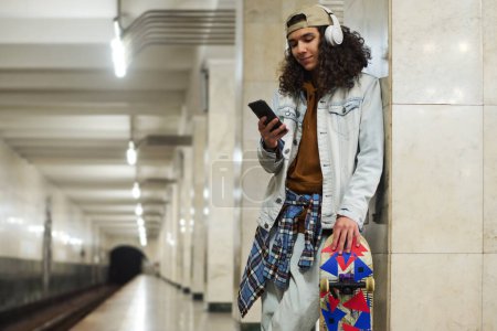 Photo for Cute smiling teenager in headphones watching online video in mobile phone while standing at subway station and waiting for train - Royalty Free Image