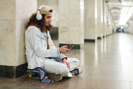 Photo for Adolescent guy in casualwear sitting on skateboard by white marble column in subway tunnel and listening to music while waiting for train - Royalty Free Image