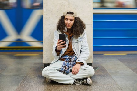 Photo for Youthul guy sitting on skateboard by marble column at subway station and communicating in video chat while looking at smartphone screen - Royalty Free Image