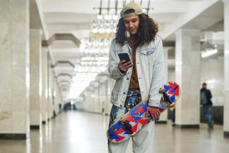 Foto de Adolescente con monopatín mirando a la pantalla del teléfono inteligente mientras camina hacia el tren subterráneo a lo largo del túnel con filas de columnas de mármol blanco - Imagen libre de derechos