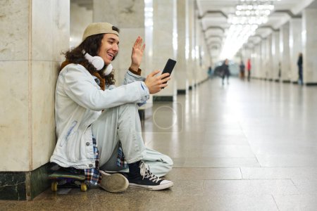 Photo for Happy teenage boy waving hand to his friend on smartphone screen while sitting on skateboard in subway tunnel and communicating in video chat - Royalty Free Image