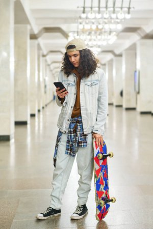 Photo for Serious teenage boy with skateboard standing at subway station and texting in smartphone while waiting for train in tunnel with columns - Royalty Free Image